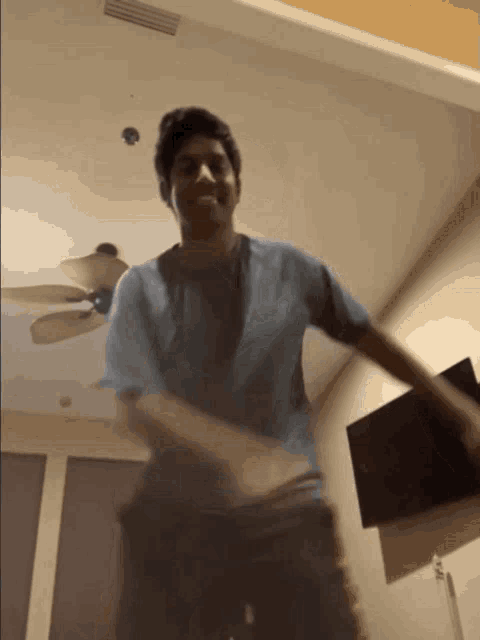 a young man in a blue shirt is dancing in a room with a ceiling fan