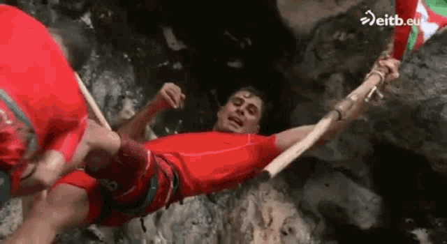 a man in a red shirt is hanging upside down from a wooden stick ..