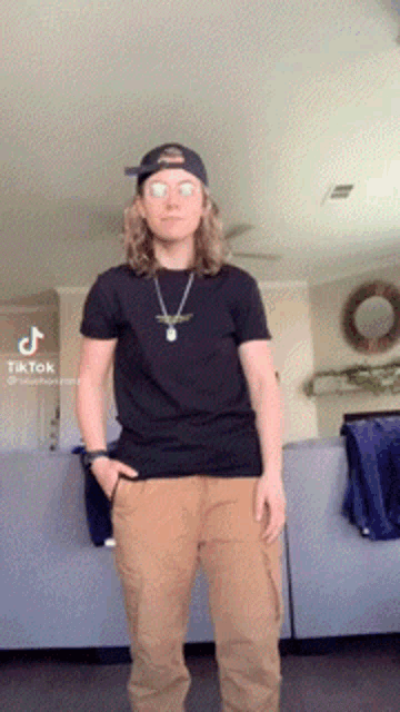a young man wearing a black shirt and brown pants is standing in a living room with his hands in his pockets ..