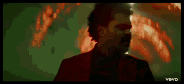 a man in a red suit and black shirt is standing in front of a fire in a dark room .