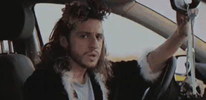 a man with long hair and a beard is driving a car while holding a pair of scissors .