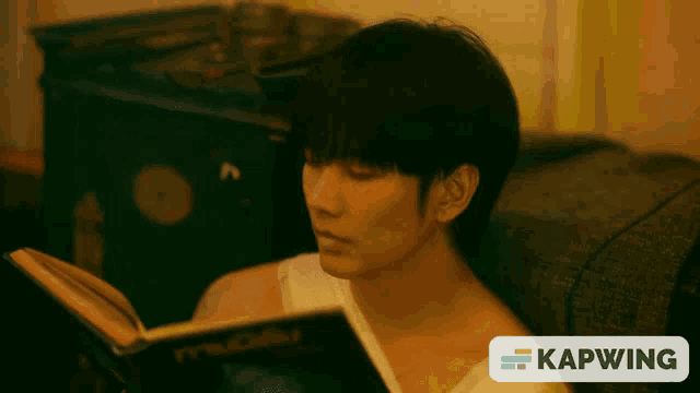 a man sits on a couch reading a book with a kapwing logo on the bottom right