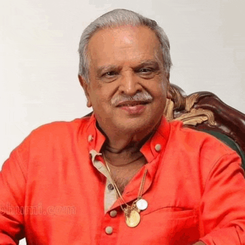 an older man wearing a red shirt and a gold necklace is smiling while sitting in a chair .