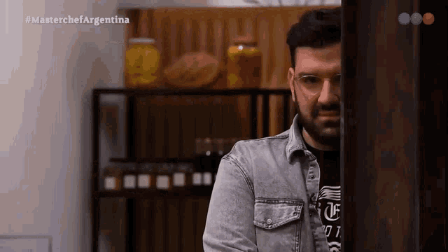 a man wearing glasses and a denim shirt is standing in front of a shelf with jars on it ..