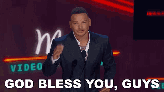 a man speaking into a microphone with the words god bless you guys behind him