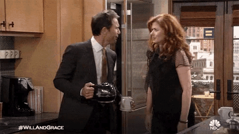 a man in a suit and tie is holding a coffee pot and talking to a woman in a kitchen .