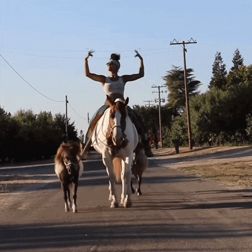 a woman riding a horse on a road with a pony behind her