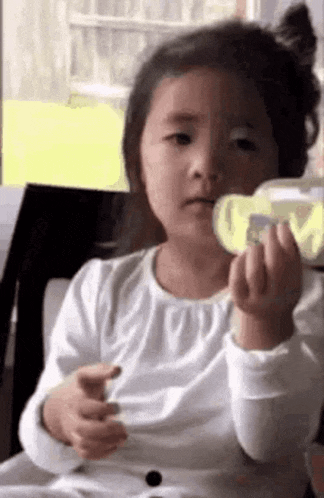 a little girl in a white shirt is sitting in a chair holding a bottle .