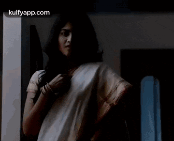 a woman in a white saree is standing in a dark room and looking at the camera .