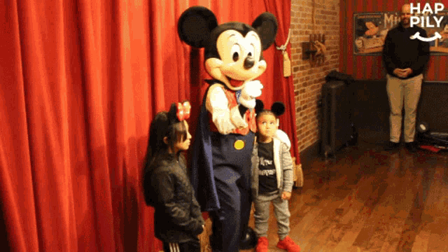 a little girl stands next to a mickey mouse mascot in front of a red curtain that says hap pily