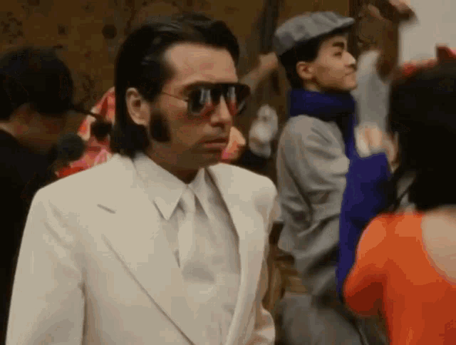 a man wearing sunglasses and a white suit stands in a crowd of people