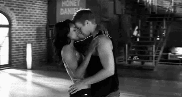 a black and white photo of a man and a woman kissing in a dance studio .