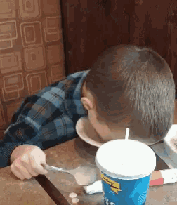 a boy is sitting at a table with a spoon in his mouth and a cup of soda