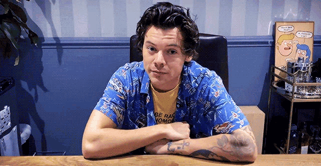 harry styles is sitting at a table with his arms crossed and wearing a blue shirt that says be raw momo .
