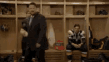 a man in a suit stands in a locker room with a hockey player sitting on a bench