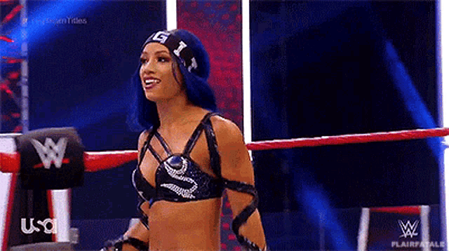 a woman with blue hair is standing in a wrestling ring wearing a black top .