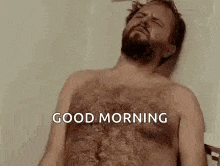a shirtless man with a beard is laying on a bed with his eyes closed and says `` good morning '' .