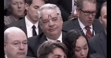 a man wearing glasses is sitting in a crowd of people in suits and ties .