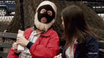 a man wearing a mask and a beard is eating a sandwich while sitting on a bench with a woman .