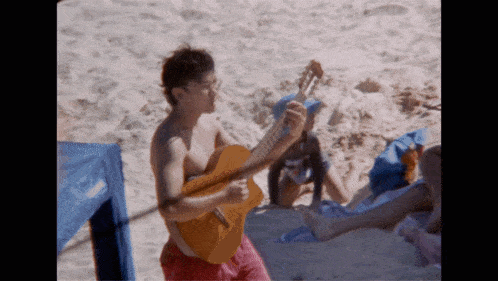 a man playing a guitar on the beach with a woman laying on the sand behind him