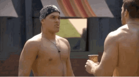 two shirtless men are standing next to each other and talking . one of the men has a bandana on his head .