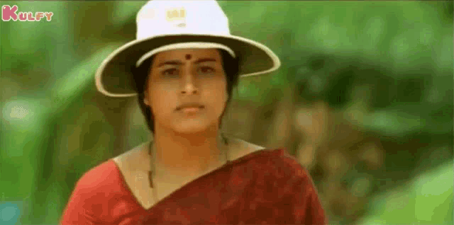 a woman wearing a white hat and a red saree is standing in the woods .