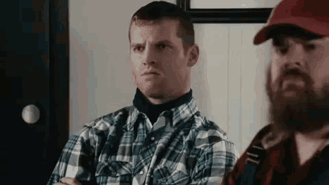 a man with a beard is sitting next to another man in a plaid shirt and hat .