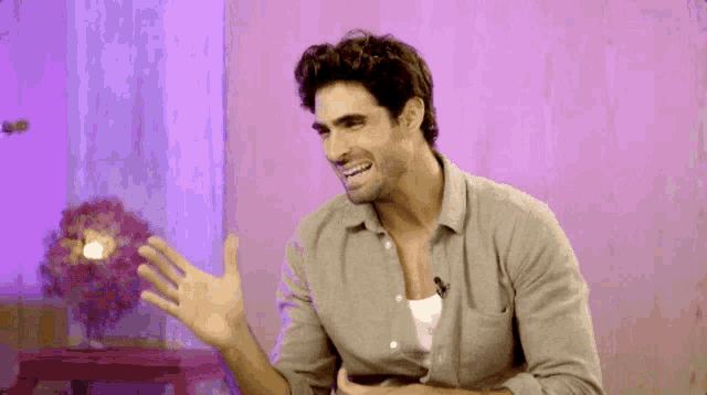 a man in a grey shirt is sitting in front of a pink wall and a purple curtain .