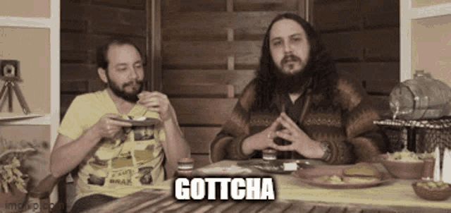 two men are sitting at a table eating food and one of them says gottcha