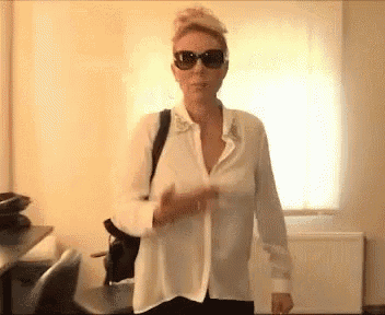 a woman wearing sunglasses and a white shirt stands in front of a window