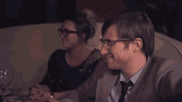 a man in a suit and tie is smiling while sitting next to a woman wearing glasses