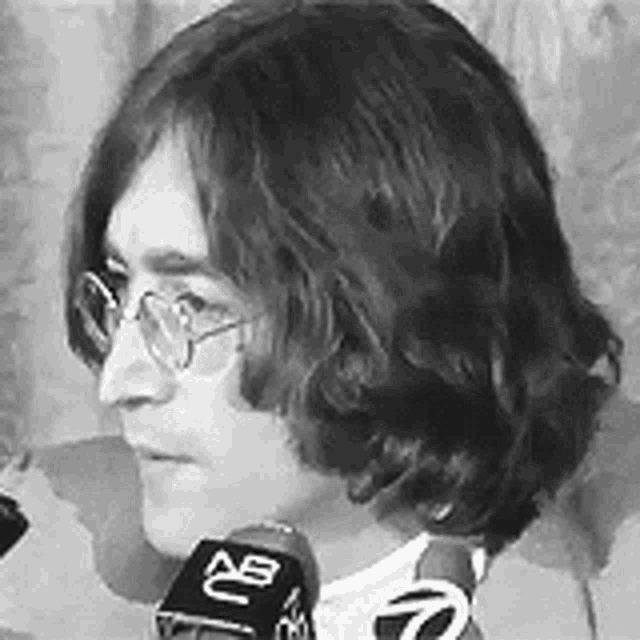john lennon is wearing glasses and talking into a microphone .