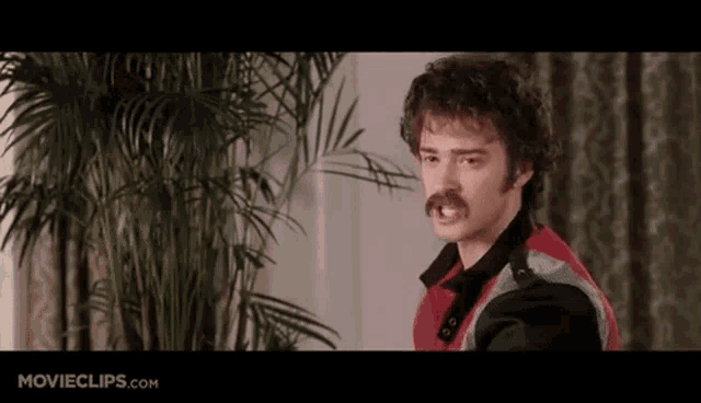 a man with a mustache is standing in front of a plant with movieclips.com written on the bottom of the screen