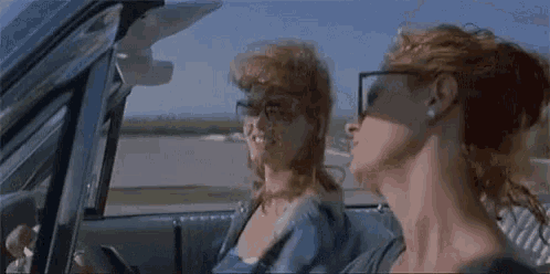 two women are sitting in a car wearing sunglasses and looking out the window .
