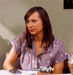 a woman in a purple shirt is sitting at a desk with what2itytv written on the bottom of her shirt .