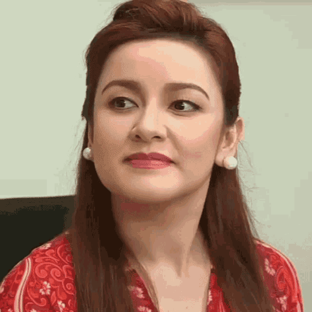 a woman with red hair and white earrings is wearing a red shirt and making a funny face .