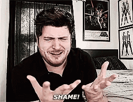 a man with a beard is sitting on a bed making a funny face and saying `` shame '' .