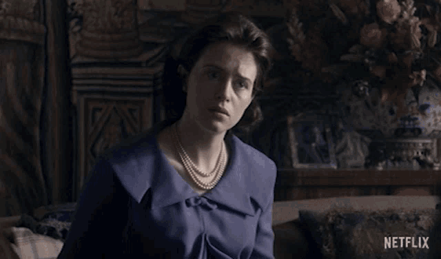 a woman with a pearl necklace is sitting on a couch and asking what