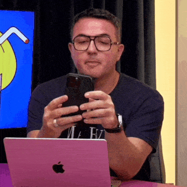 a man wearing glasses looks at his cell phone while sitting in front of an apple laptop