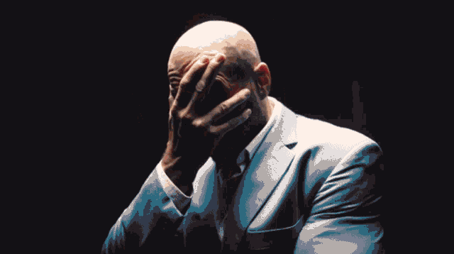 a bald man in a blue suit covering his face with his hands