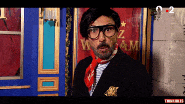a man wearing glasses and a red scarf stands in front of a sign that says " waz am "