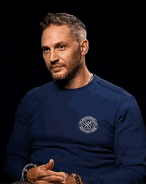 a man wearing a blue sweater is laughing while sitting down with his hands folded .