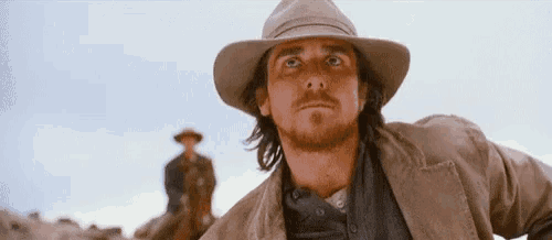 a man wearing a hat and a jacket is standing in front of a man on a horse .