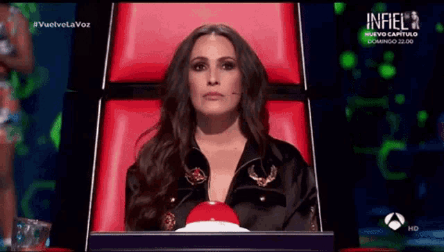 a woman sits in a chair with a red button in front of her and the word infiel is on the screen behind her