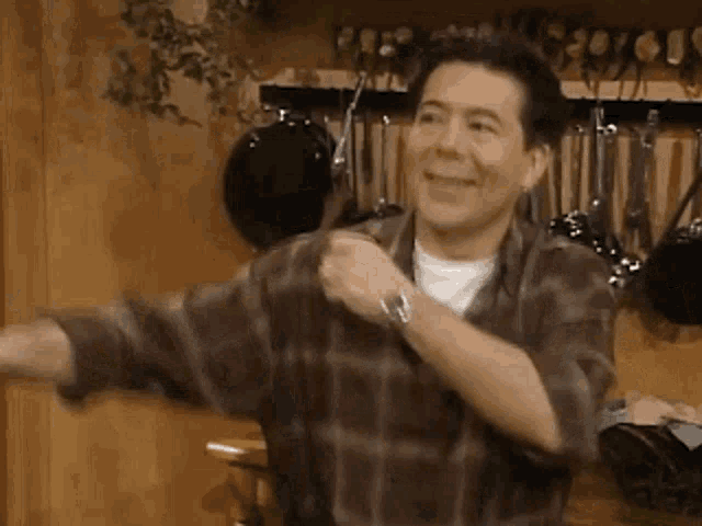 a man in a plaid shirt is dancing in a kitchen with pots and pans hanging on the wall behind him