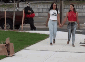 two women are walking down a sidewalk holding hands while a man in a mask is walking behind them .