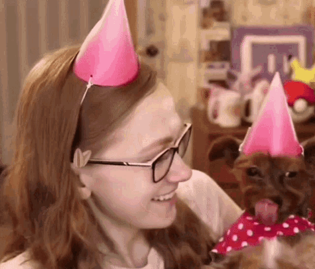 a woman wearing glasses and a pink party hat looks at a dog wearing a pink party hat