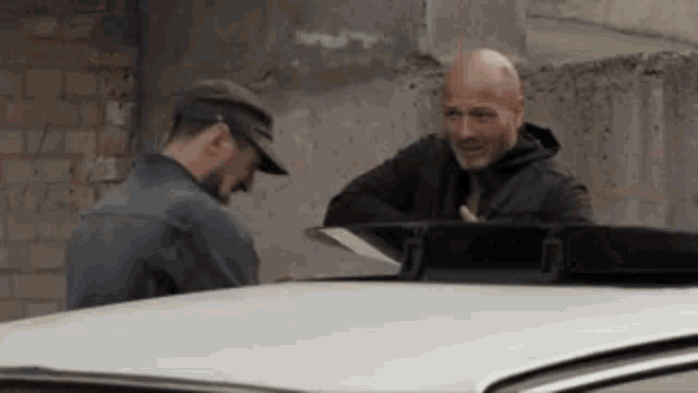 two men are standing next to a car and talking .