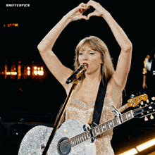 a woman is singing into a microphone while holding a guitar and making a heart shape with her hands