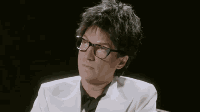 a man with glasses and a white jacket is sitting in a chair .